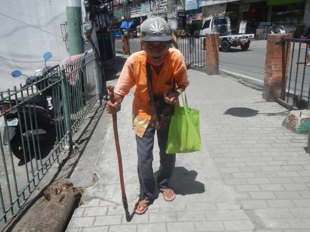 Ein älterer Mann geht die Straße entlang, in einer Hand einen Stock, in der anderen eine Tasche und einen Stock, trägt eine Mütze, mit einem Metallzaun links und Fahrzeugen, Gebäuden und Schildern im Hintergrund.