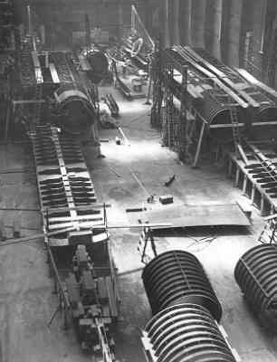 Schwarzes und weißes Foto eines industriellen Fabrikinneren mit umfangreichen Rohrleitungen, Maschinen und Geräten auf dem Boden, das strukturelle Pfeiler und eine entfernte Wand zeigt.