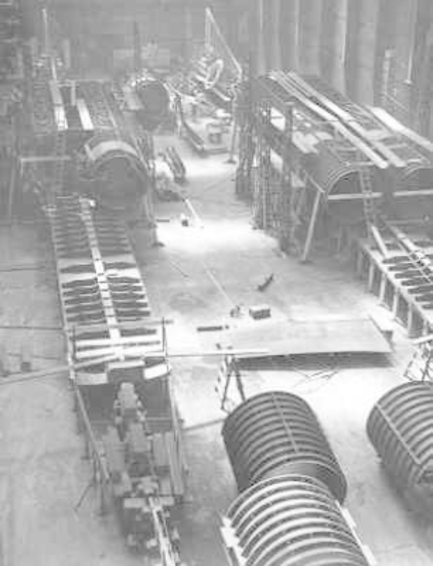 Schwarzes und weißes Foto eines industriellen Fabrikinneren mit umfangreichen Rohrleitungen, Maschinen und Geräten auf dem Boden, das strukturelle Pfeiler und eine entfernte Wand zeigt.