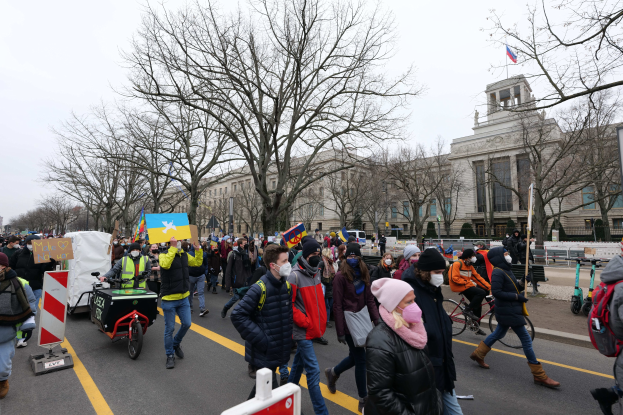 Eine große Gruppe von Menschen marschiert auf einer Stadtstraße bei einer Demonstration, einige halten Schilder und andere fahren Fahrräder, mit Bäumen und einem Gebäude im Hintergrund unter einem klaren blauen Himmel.