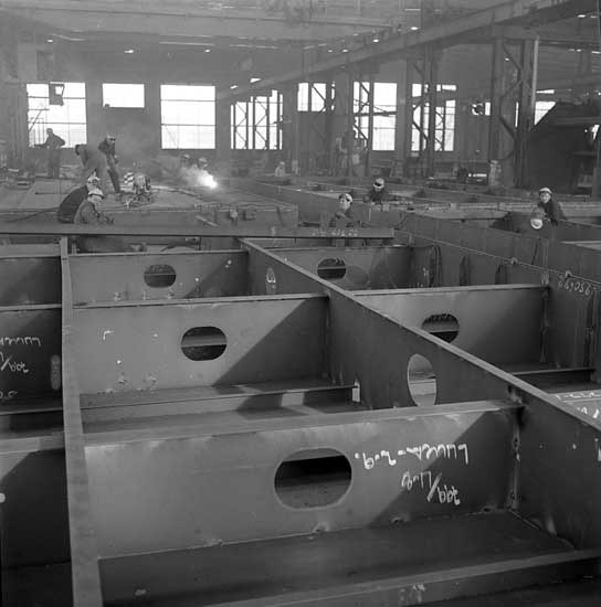 Schwarzes und weißes Foto einer Fabrik, in der Arbeiter in Helmen Stahlträger zusammenbauen, mit Fenstern, Pfählen, Lampen und Text im Hintergrund.