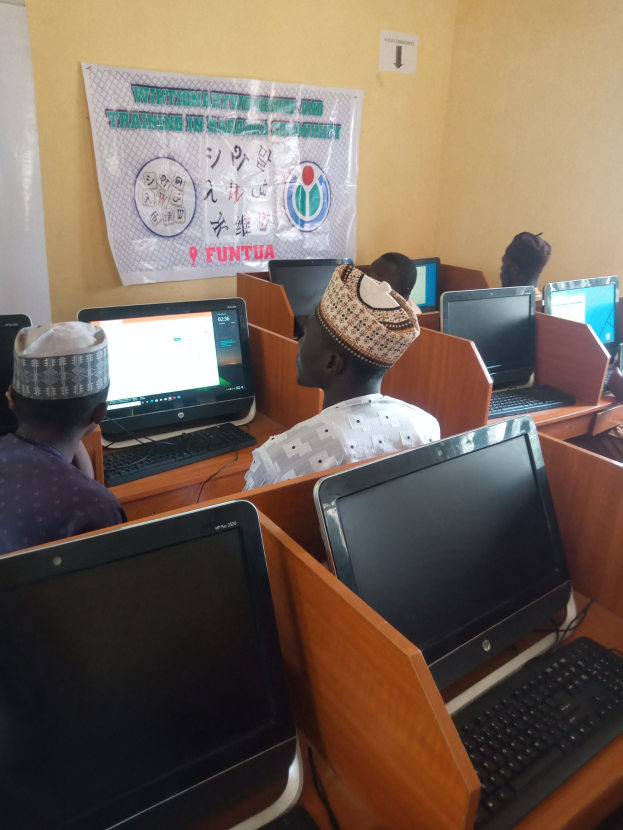 Eine Gruppe von Menschen, die an Tischen mit Computern, Monitoren, Tastaturen und Mäusen sitzen und an einem Workshop zur Computertechnologie teilnehmen, mit einem Plakat im Hintergrund.