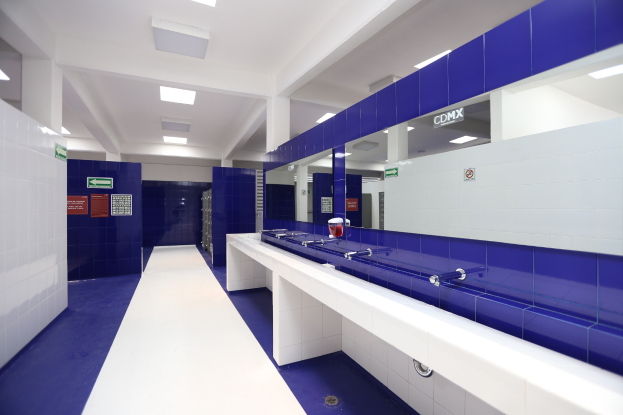 Ein Öffentliches WC mit blauen Fliesenwänden und weißen Arbeitsplätzen, mit Waschbecken mit Hähnen, einer Plattform mit einer Flasche und anderen Gegenständen, Texttafeln an den Wänden, Deckenleuchten und Pfeilern.