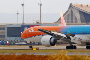 KLM Boeing 777-300ER auf dem Frankfurter Flughafen-Ring mit Gras und Pflanzen im Vordergrund, Gebäuden und Fahrzeugen im Hintergrund unter einem klaren blauen Himmel.