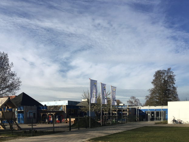Großes Gebäude mit einem grasigen Bereich davor, umgeben von einem Metallzaun, mit einem Weg, Bäumen, Pfosten, Bannern, einem geparkten Fahrrad, nahegelegenen Häusern und einem bewölkten Himmel, als neuer Spielbereich für Kinder an der Schule identifiziert.