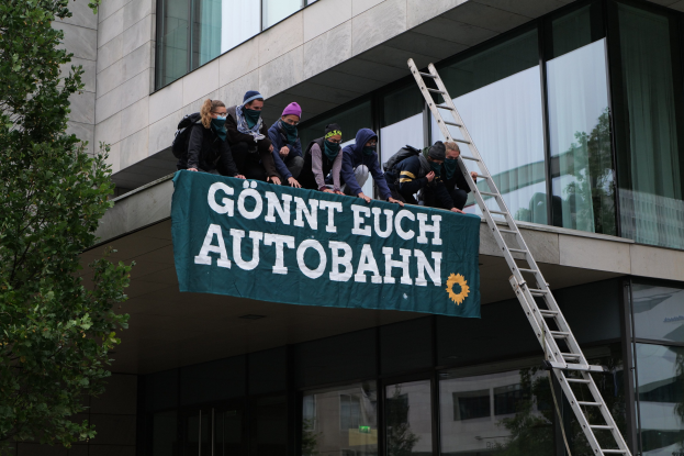 Menschen auf einem Gebäude mit einer Fahne, einem Baum auf der linken Seite und Glasfenstern im Hintergrund, wahrscheinlich protestieren sie für die deutsche Regierung.