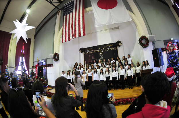 Eine Gruppe von Menschen auf einer Bühne mit Weihnachtsdekoration, einem Rednerpult, Lautsprechern, einer Texttafel und einer Menge mit Kameras und Handys.