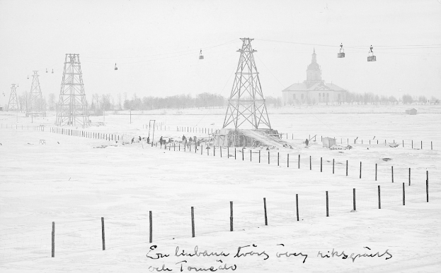 Schwarze und weiße Aufnahme einer Skihebeanlage in einem verschneiten Feld mit Stützpfählen, Überseilbahn, Bäumen und einem Gebäude im Hintergrund, mit Text am unteren Rand.