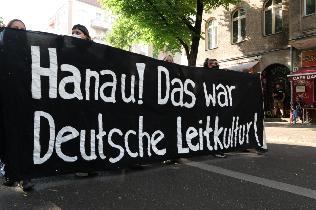 Eine Gruppe von Menschen, die eine Stra√e entlanggehen und eine schwarze Fahne mit der Aufschrift "Hanau das war deutsche Leitkultur" tragen, mit Gebäuden, Bäumen und einem klaren blauen Himmel im Hintergrund.
