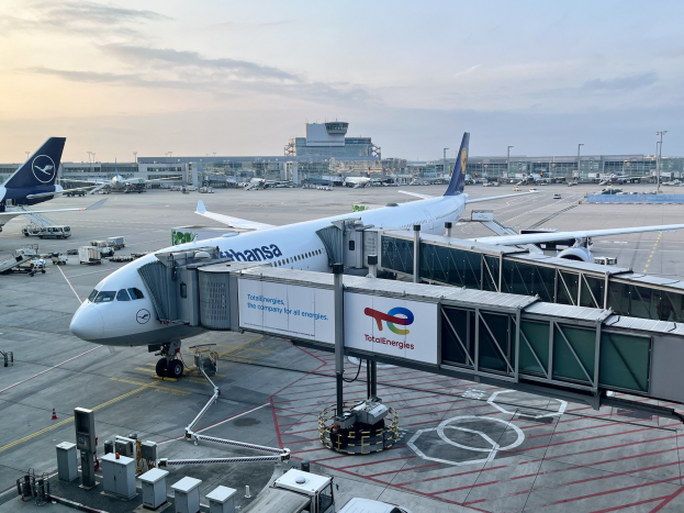 Großraumjet auf einem Flughafenvorfeld mit anderen Flugzeugen, Fahrzeugen und Flughafeninfrastruktur im Hintergrund unter einem klaren blauen Himmel.