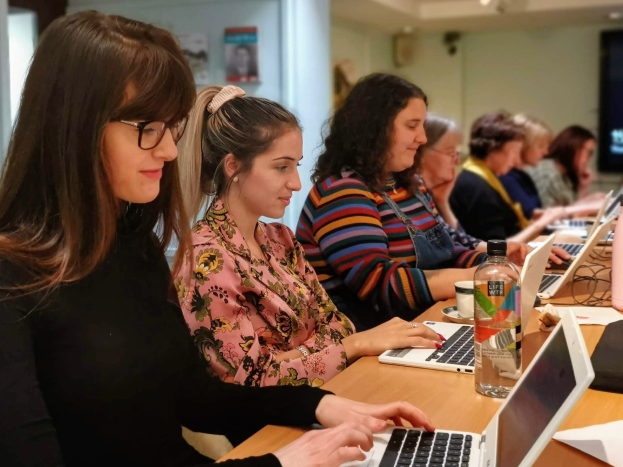 Eine Gruppe von Frauen, die um einen Tisch herum auf Laptops arbeiten, in einem Raum mit einem Foto Rahmen, einer Säule und Deckenleuchten, wahrscheinlich an einem Online-Kurs teilnehmen.