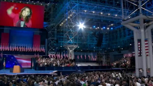 Eine große Menschenmenge sitzt vor einer Bühne mit einem Rednerpult, Mikrofon, Flaggen, einem Bildschirm mit dem Bild einer Frau und Beleuchtungsausrüstung, wahrscheinlich bei einer Präsidenten-Debatte.