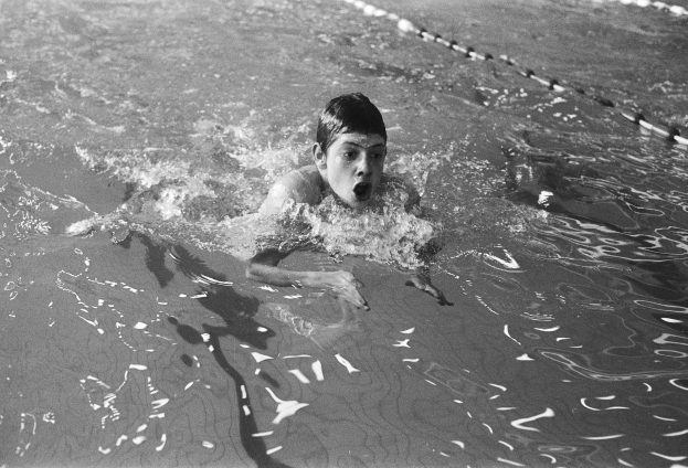 Schwarzes und weißes Foto eines jungen Jungen, der im Pool schwimmt und den Mund öffnet, umgeben von Schwimmbahnen.