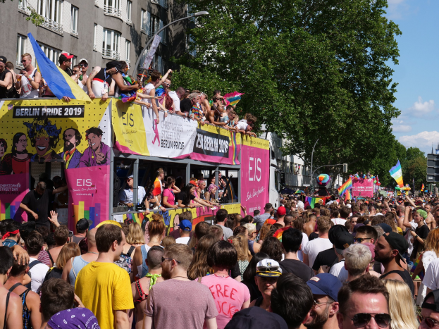 Eine große Menge marschiert während des Berlin Pride 2019 die Straße entlang, viele tragen Kappen und Goggles, einige halten Fahnen, mit Bannern, Laternenmasten, Bäumen und Gebäuden im Hintergrund unter einem bewölkten Himmel.