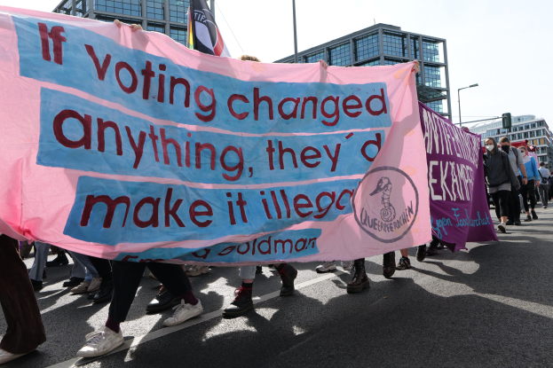 Eine Gruppe von Menschen marschiert die Straße entlang und trägt ein Banner mit der Aufschrift "Wenn Abstimmen etwas ändern würde, hätten sie es verboten", mit Gebäuden, Laternenpfählen und einem klaren blauen Himmel im Hintergrund.
