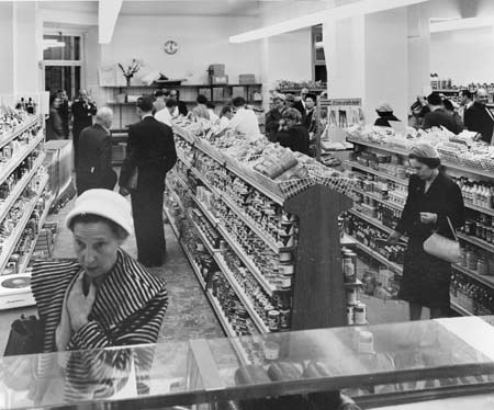 Schwarzes und weißes Foto von Einkäufern in einem Supermarkt mit Lebensmittelregalen, Taschen, einer Wanduhr und Deckenbeleuchtung.