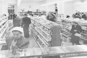 Schwarzes und weißes Foto von Einkäufern in einem Supermarkt mit Lebensmittelregalen, Taschen, einer Wanduhr und Deckenbeleuchtung.