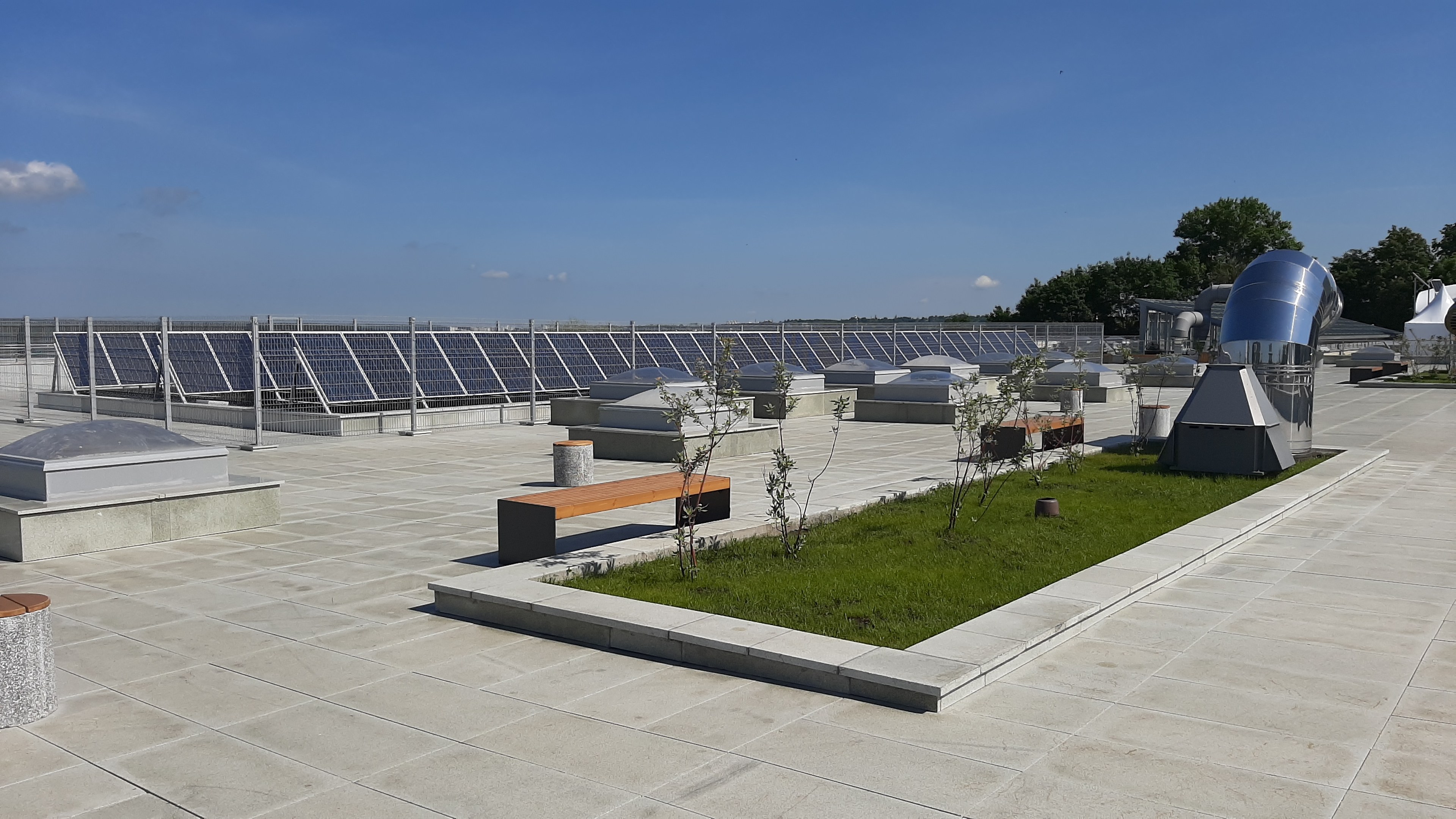 Ein Gebäude mit Solarmodulen auf dem Dach, umgeben von Bänken, Pflanzen, Gras und Bäumen unter einem bewölkten Himmel.