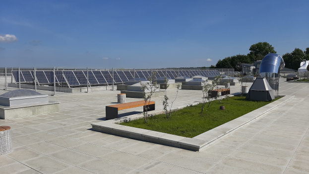 Ein Gebäude mit Solarmodulen auf dem Dach, umgeben von Bänken, Pflanzen, Gras und Bäumen unter einem bewölkten Himmel.