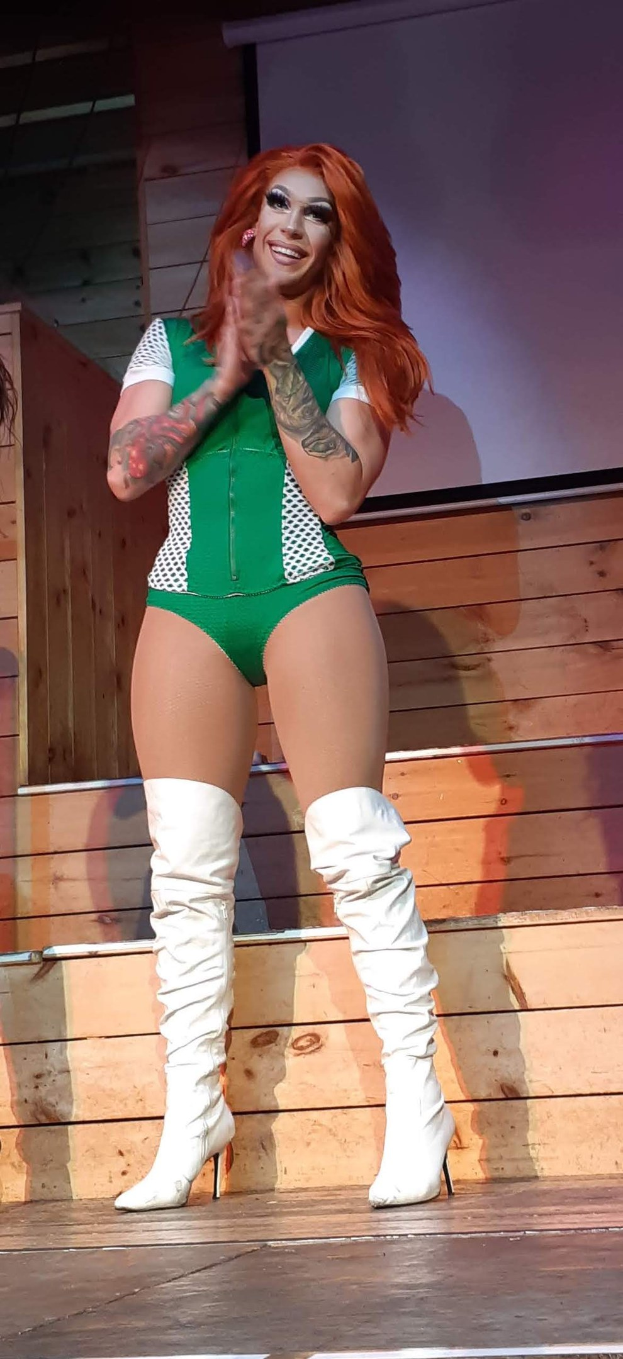 Frau in grüner Bodysuit und weißen Stiefeln auf einer Bühne, die wie eine Wrestlerin aussieht, mit einer hölzernen Wand und einem Hintergrundbild.
