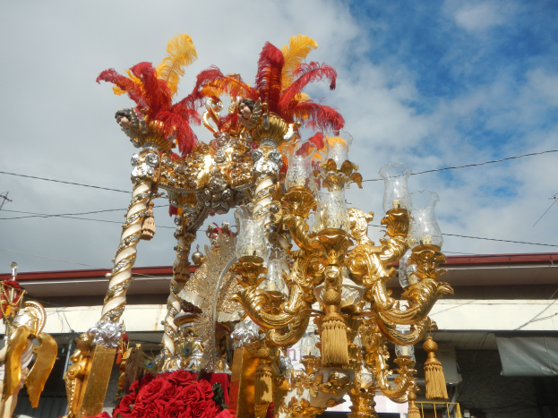 Ein großes goldenes und rotes Festwagen mit Blumen und anderen Schmuckelementen bei einem Karnevalsumzug, mit einem Gebäude, Strommasten mit Drähten und einem bewölkten Himmel im Hintergrund.