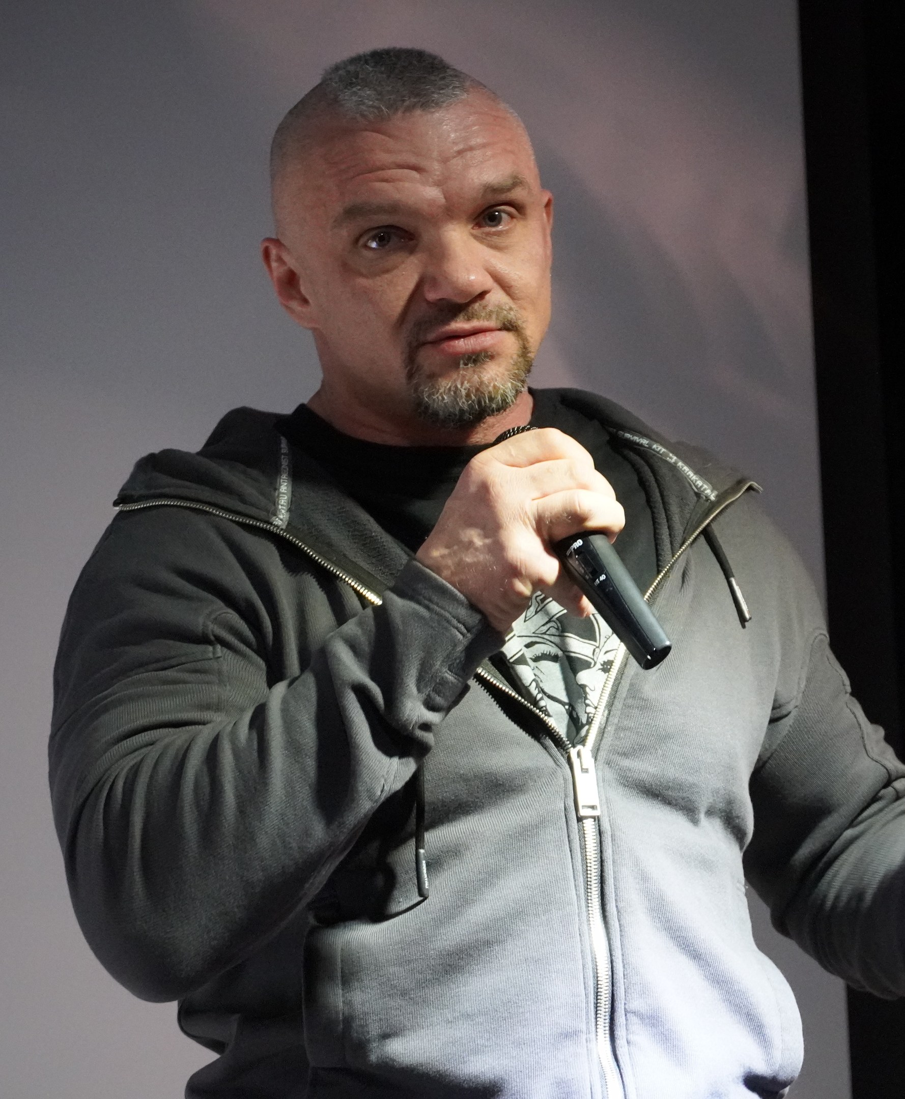 Ein Mann in einer schwarzen Kapuzenjacke und einem schwarzen Anzug mit einem weißen Hemd, zurückgekämmtem Haar und einem selbstbewussten Gesichtsausdruck hält ein Mikrofon, während er vor einer Wand steht.