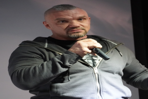 Ein Mann in einer schwarzen Kapuzenjacke und einem schwarzen Anzug mit einem weißen Hemd, zurückgekämmtem Haar und einem selbstbewussten Gesichtsausdruck hält ein Mikrofon, während er vor einer Wand steht.