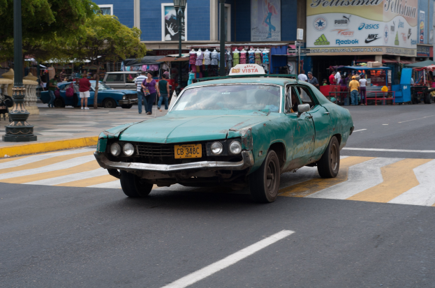 Ein grünes Auto fährt auf einer Straße mit hohen Gebäuden, Fußgängern auf dem Gehweg, Laternenpfählen und Bäumen, mit Ständen und Schildern im Hintergrund.