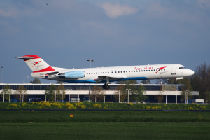 Ein Airbus A320-200 der Austrian Airlines startet am Frankfurter Flughafen, umgeben von grünem Gras, blühenden Pflanzen, Bäumen, Laternenmasten und einem Gebäude im Hintergrund bei bewölktem Himmel.