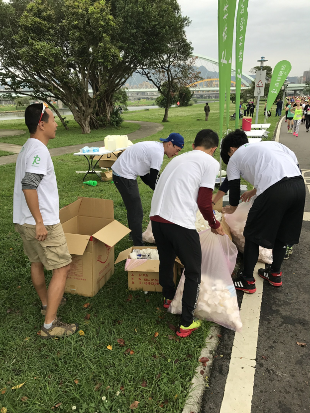 Group of people on a roadside with scattered cardboard boxes, bottles, and banners, participating in a charity run with trees, buildings, and a clear blue sky in the background.