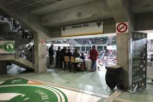Gruppe von Menschen vor einer sitzenden Menge in einem Stadion mit Schildern, die "Boston Celtics vs New York Jets" lesen, einem Müllcontainer, einer Tafel mit Text, einer Treppe mit Geländern und einer Decke mit Deckenleuchten.