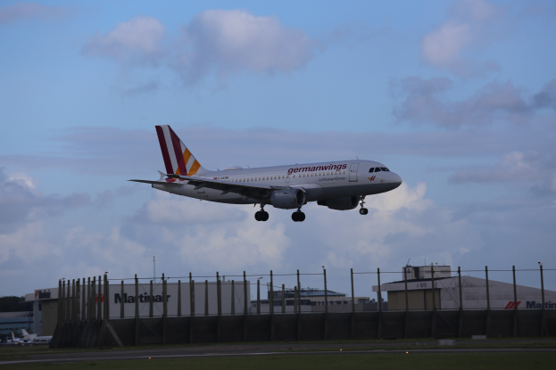 Germanwings Airbus A320-200 beim Start vom Frankfurter Airport mit Gras darunter, einem Zaun im Vordergrund, Gebäuden und Bäumen im Hintergrund und einer bewölkten Himmel