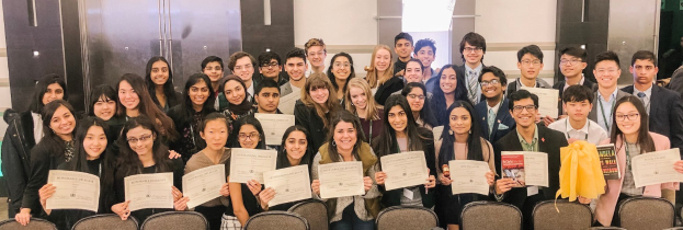 Gruppe von Menschen mit Urkunden, die zusammen in einem Raum mit Stühlen und einer Wand im Hintergrund stehen, wahrscheinlich feiern sie eine Abschlussfeier für das Studienjahr 2019-2020.