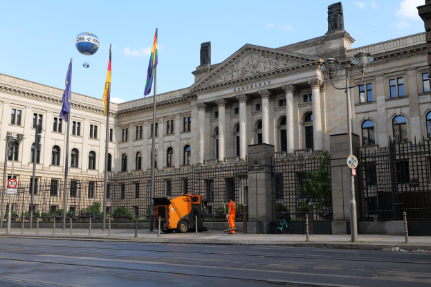 Das Reichstagsgebäude in Berlin, Deutschland, mit vielen Fenstern, umgeben von Straßenlaternen, Schildern, Fahrzeugen, Menschen, Flaggen, Gittern und Bäumen, unter einem bewölkten Himmel.
