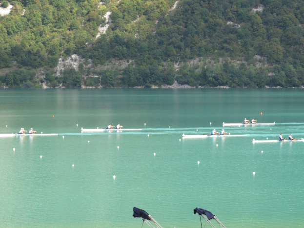 Eine Gruppe von Menschen, die auf einem Gewässer umgeben von Bäumen und einem Hügel rudern, mit Bekleidungsständern am Boden, die auf einen Ruderwettbewerb hinweisen.