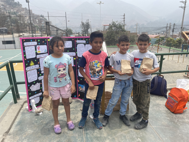Gruppe von Kindern auf einem Basketballfeld mit Papiertüten in den Händen, mit Tüten und Gegenständen rechts daneben und einer Tafel mit Text links daneben, vor dem Hintergrund von Zäunen, Pfosten, Straßenlaternen, Bäumen, Gebäuden, Hügeln und dem Himmel.