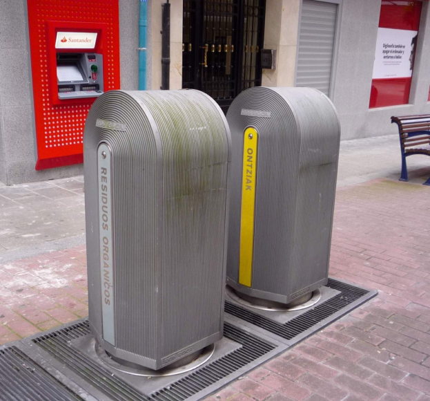 Zwei Metallmülltonnen auf einem Bürgersteig neben einer Bank und einem Gebäude, mit einem Geldautomaten links im Bild.