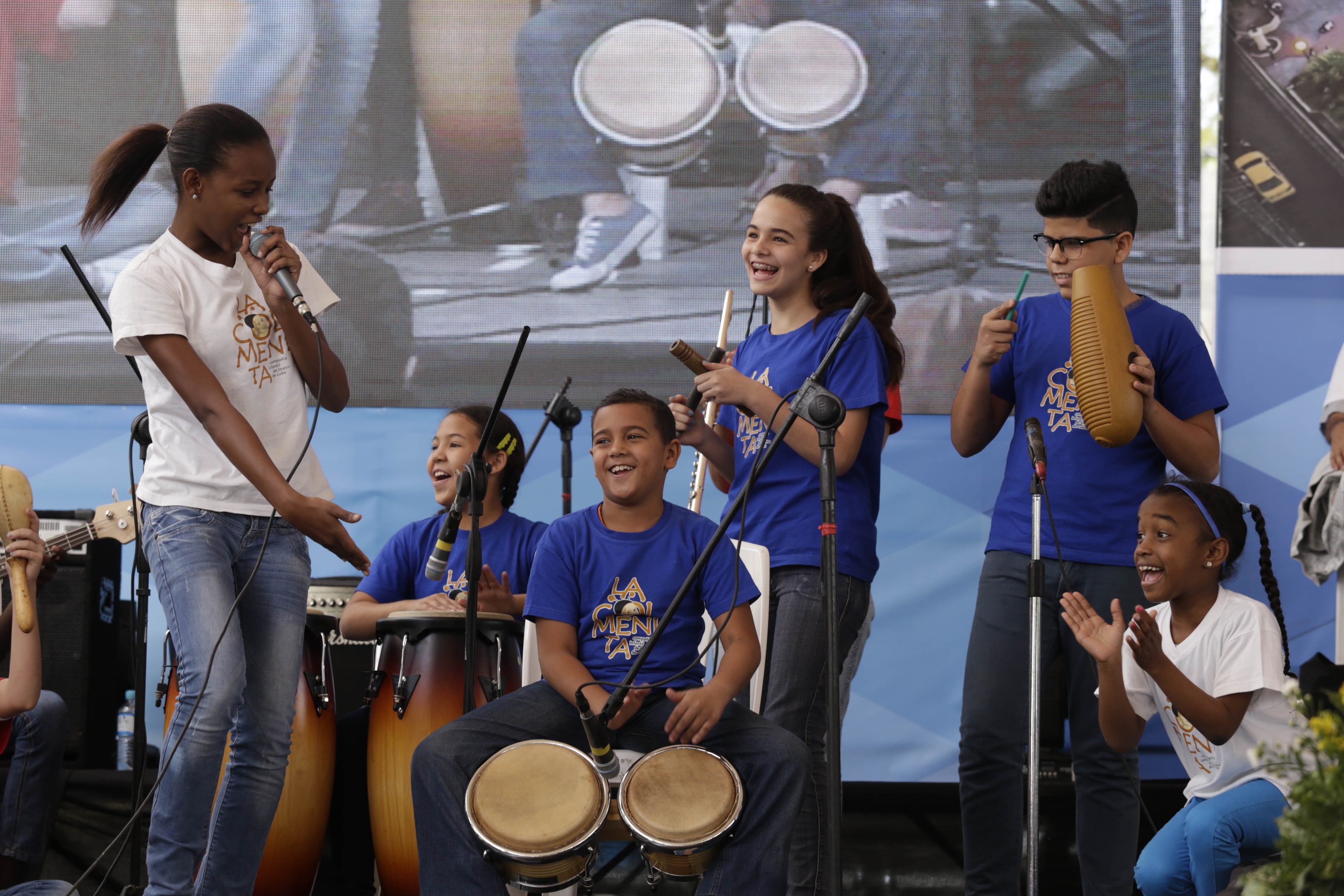 Eine Gruppe von Kindern, die auf einer Bühne Musikinstrumente spielen, mit einer Frau, die ein Mikrofon hält, in der Mitte, umgeben von Mikrofonständern, einem Banner im Hintergrund und Pflanzen auf der rechten Seite.