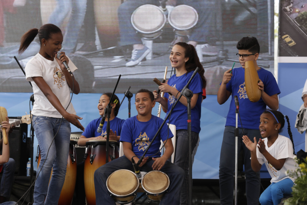 Eine Gruppe von Kindern, die auf einer Bühne Musikinstrumente spielen, mit einer Frau, die ein Mikrofon hält, in der Mitte, umgeben von Mikrofonständern, einem Banner im Hintergrund und Pflanzen auf der rechten Seite.