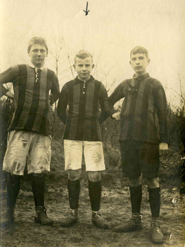 Drei junge Männer in Fußballtrikots stehen zusammen auf einem grünen Feld umgeben von Bäumen unter einem klaren Himmel.