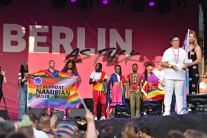 Gruppe von Menschen auf einer Bühne mit einem Banner 'Berlin as Fuck' und Lautsprechern, Mikrofonstativen und einer feiernden Menge unten, die Hände hebt.