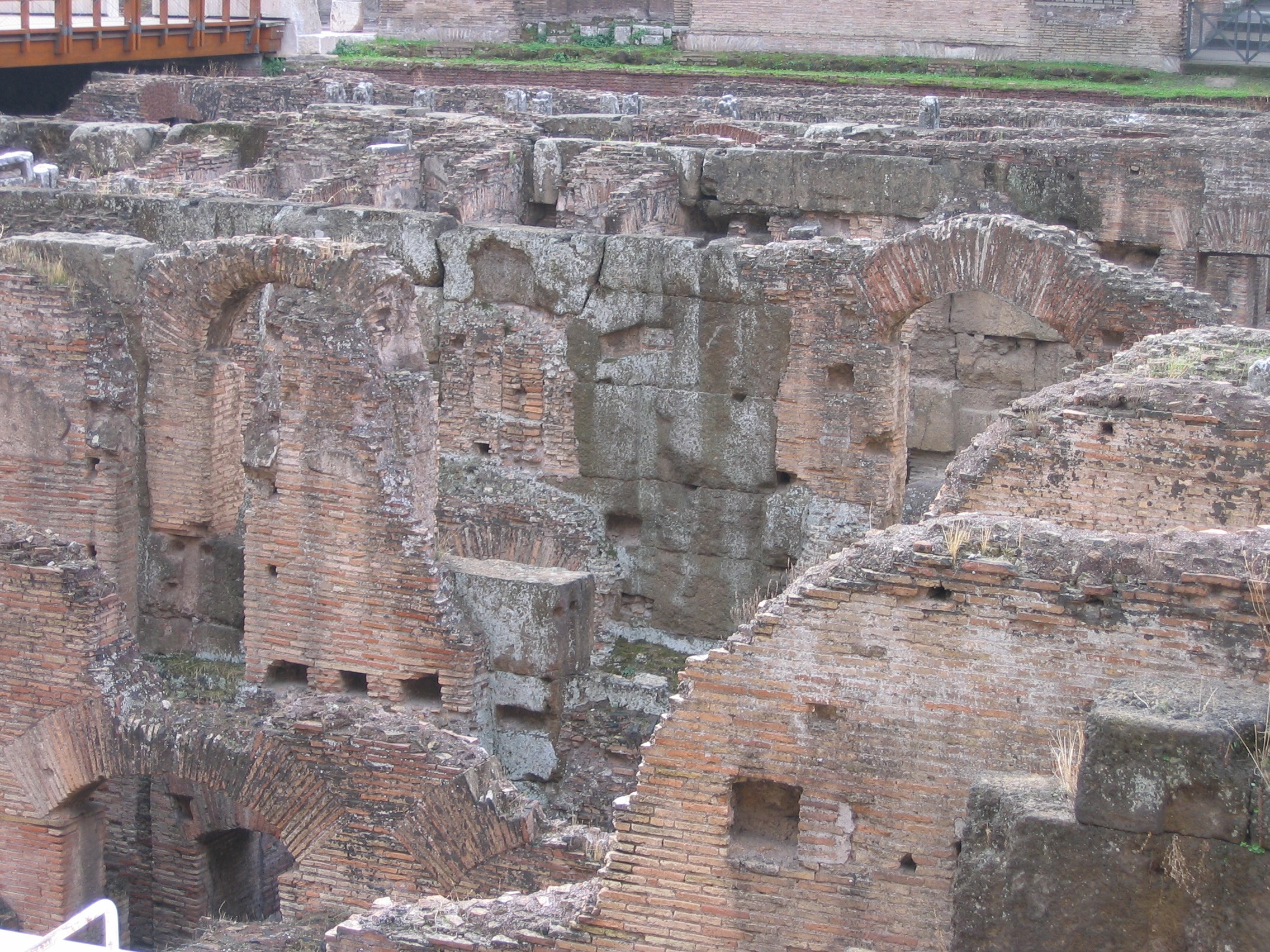 Römische Forumruinen mit verwitterten Steinwänden, Bögen und Säulen, grasbewachsenem Boden, einer hölzernen Brücke mit Geländern auf der linken Seite und fernen Gebäuden im Hintergrund.