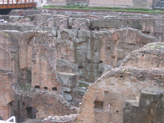 Römische Forumruinen mit verwitterten Steinwänden, Bögen und Säulen, grasbewachsenem Boden, einer hölzernen Brücke mit Geländern auf der linken Seite und fernen Gebäuden im Hintergrund.