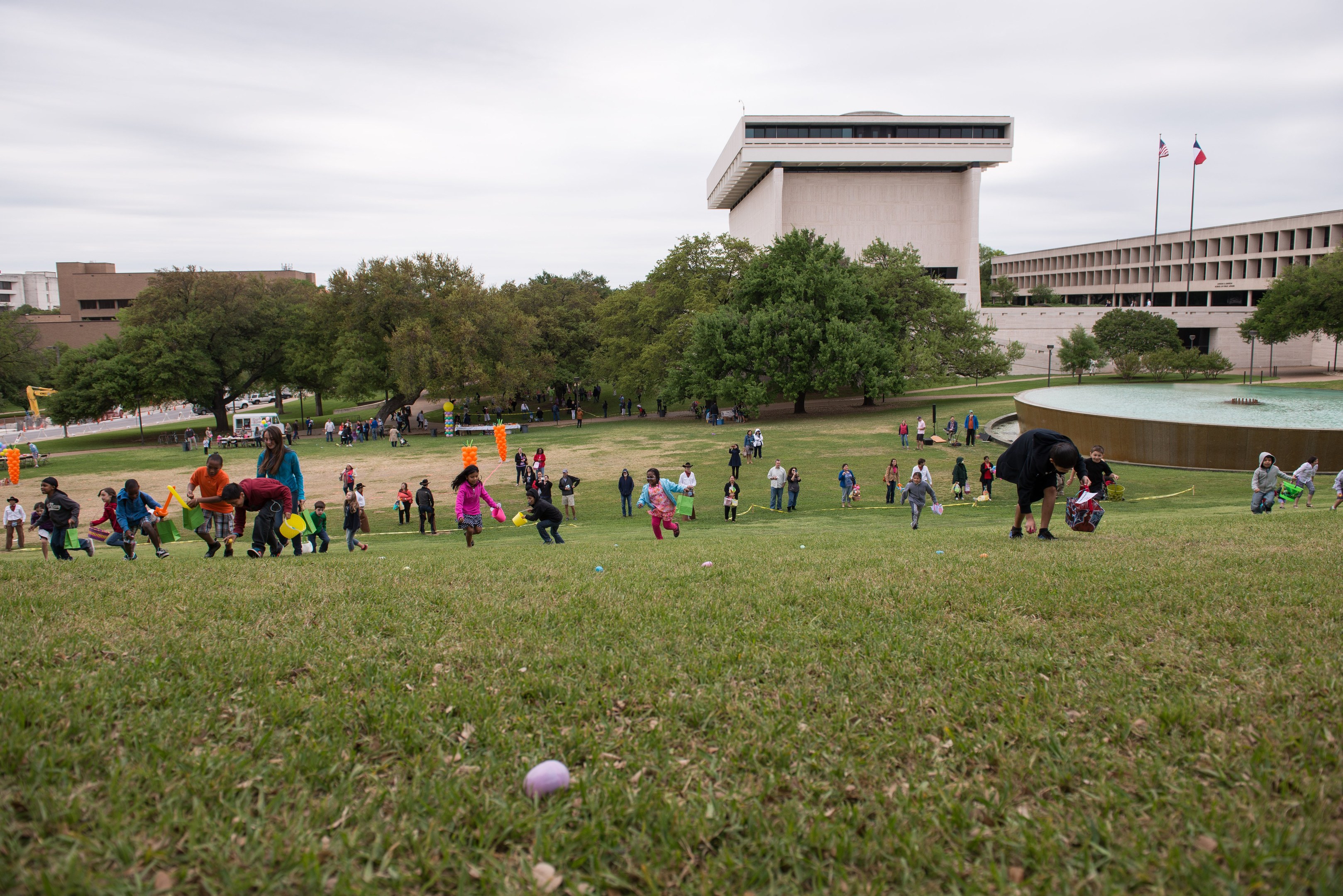 Eine Gruppe von Menschen spielt mit Ostereiern auf einer grünen Wiese mit Bäumen, Gebäuden, Flaggen, einem Brunnen und einem bewölkten Himmel im Hintergrund.