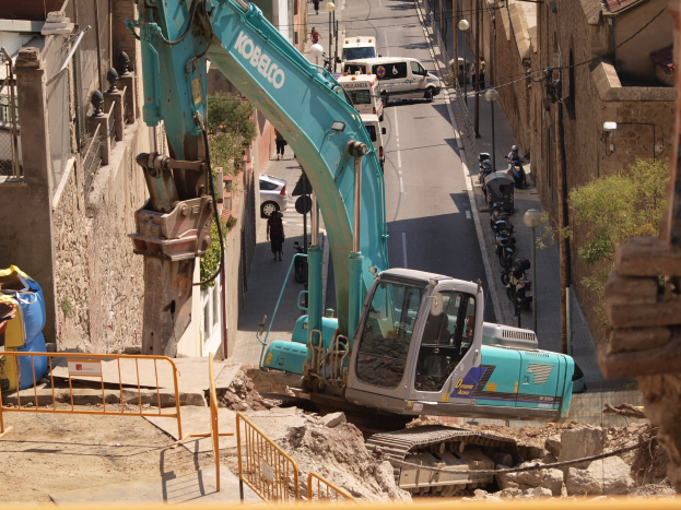 Bagger am Einsatz auf einer Baustelle mit Gebäuden, Straßeninfrastruktur, Fahrzeugen, Fußgängern und Grünflächen.