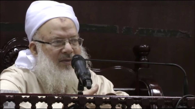 Asaduddin Owaisi, ein Mann mit einem weißen Bart und Turban, spricht in ein Mikrofon vor einem hölzernen Geländer, trägt ein cremefarbenes Gewand und Brille, vor einem dunklen Hintergrund.