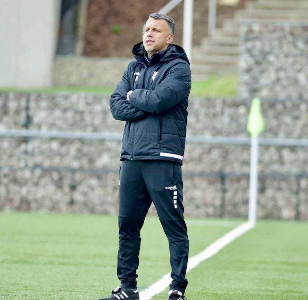Ein Fußballtrainer steht auf einem Feld und hat die Arme verschränkt, trägt eine schwarze Jacke und Hose, vor einem Zaun, Treppe und Wand während eines Trainings.