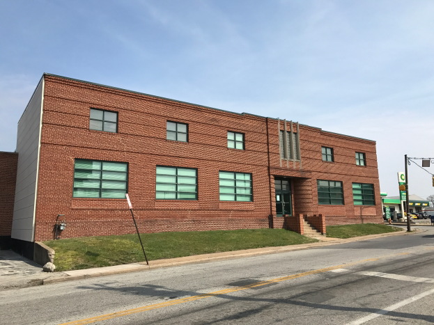 Ein großes Backsteinschulgebäude an einer Straßenecke mit Glasfenstern, Treppen, Geländern, Rasen, Laternen, Schilder, Fahrzeugen, Bäumen und einem bewölkten Himmel.