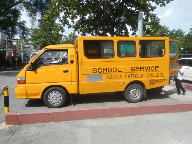Ein gelber Schulbus steht am Straßenrand geparkt, mit einer Person, die auf dem Gehweg rechts davon läuft; Bäume, Gebäude, Pfosten und ein klarer blauer Himmel sind im Hintergrund zu sehen.