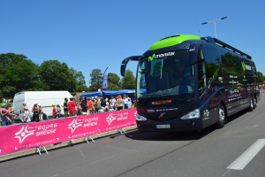 Schwarzer und grüner Bus fährt auf einer Straße neben einer Menschenmenge, einige tragen Mützen, mit einem Banner auf der linken Seite, Bäumen und einem klaren blauen Himmel im Hintergrund.